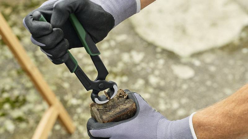 Une personne tient un sabot de cheval et utilise une pince à sabot pour le soin des sabots. Elle porte des gants gris et travaille en extérieur.
