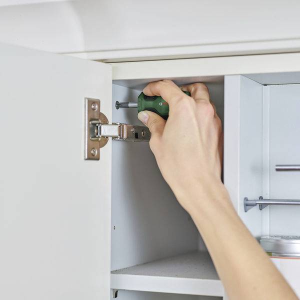 Une personne resserre une vis avec un tournevis sur une charnière de cuisine. Une armoire est légèrement entrouverte.