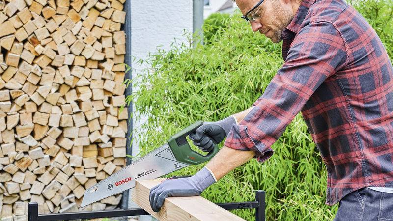 Un homme portant une chemise à carreaux scie un morceau de bois avec une scie de menuisier. Des troncs d'arbres empilés sont visibles en arrière-plan.