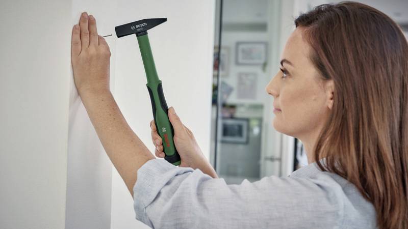 Une femme enfonce un clou dans un mur. Elle utilise un marteau à manche vert, concentrée et prudente dans son travail.