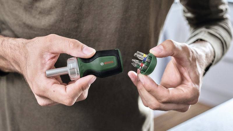 Une personne tient un tournevis Bosch vert avec une poignée amovible à capsule contenant différents embouts.