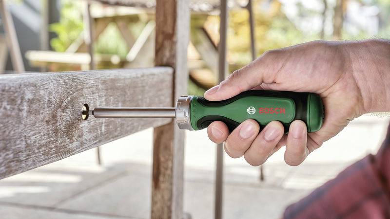 Une main utilise un tournevis Bosch vert pour serrer une vis dans un meuble en bois à l'extérieur.
