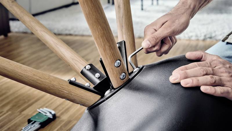Une personne fixe des pieds en bois sur un meuble rembourré à l'aide d'une clé Allen. Le sol est en bois, avec un tapis en arrière-plan.