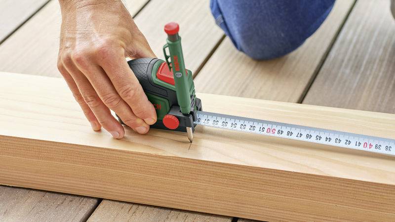 Une personne mesure une planche de bois à l'aide d'un mètre ruban fixé à un télémètre laser. La planche est posée sur des madriers en bois.
