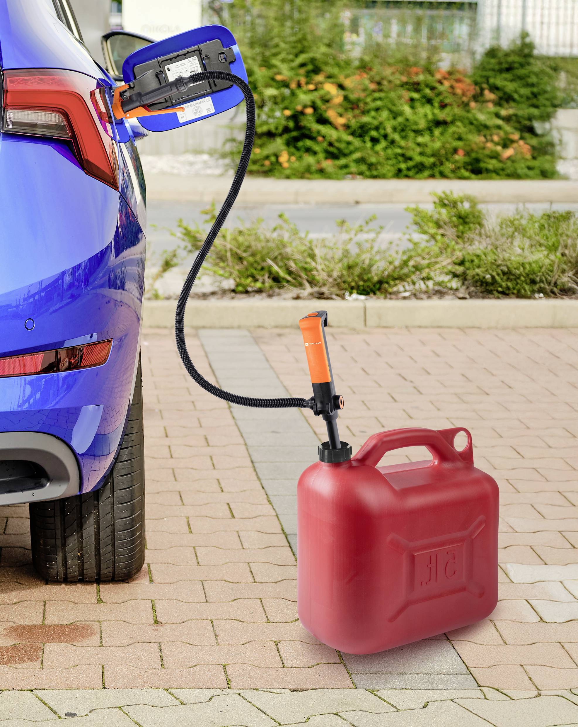 Un bidon de carburant rouge relié à une voiture par un tube, stationné sur une allée de briques avec de la verdure en arrière-plan.