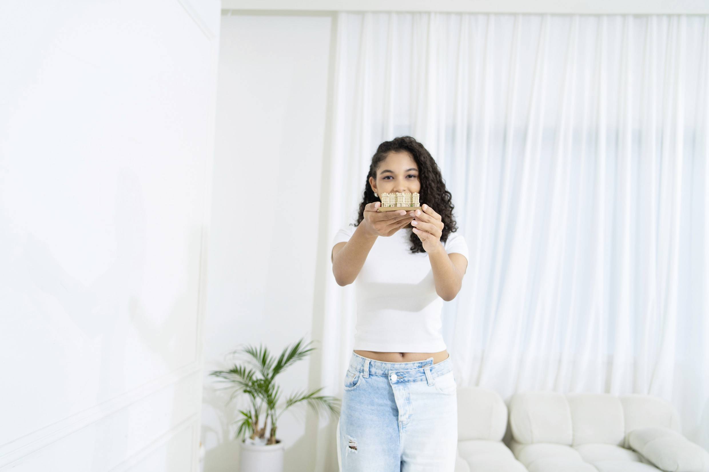 Une femme en jeans et t-shirt blanc tient un petit bloc de bois en souriant dans le salon. Des rideaux et une plante sont visibles en arrière-plan.
