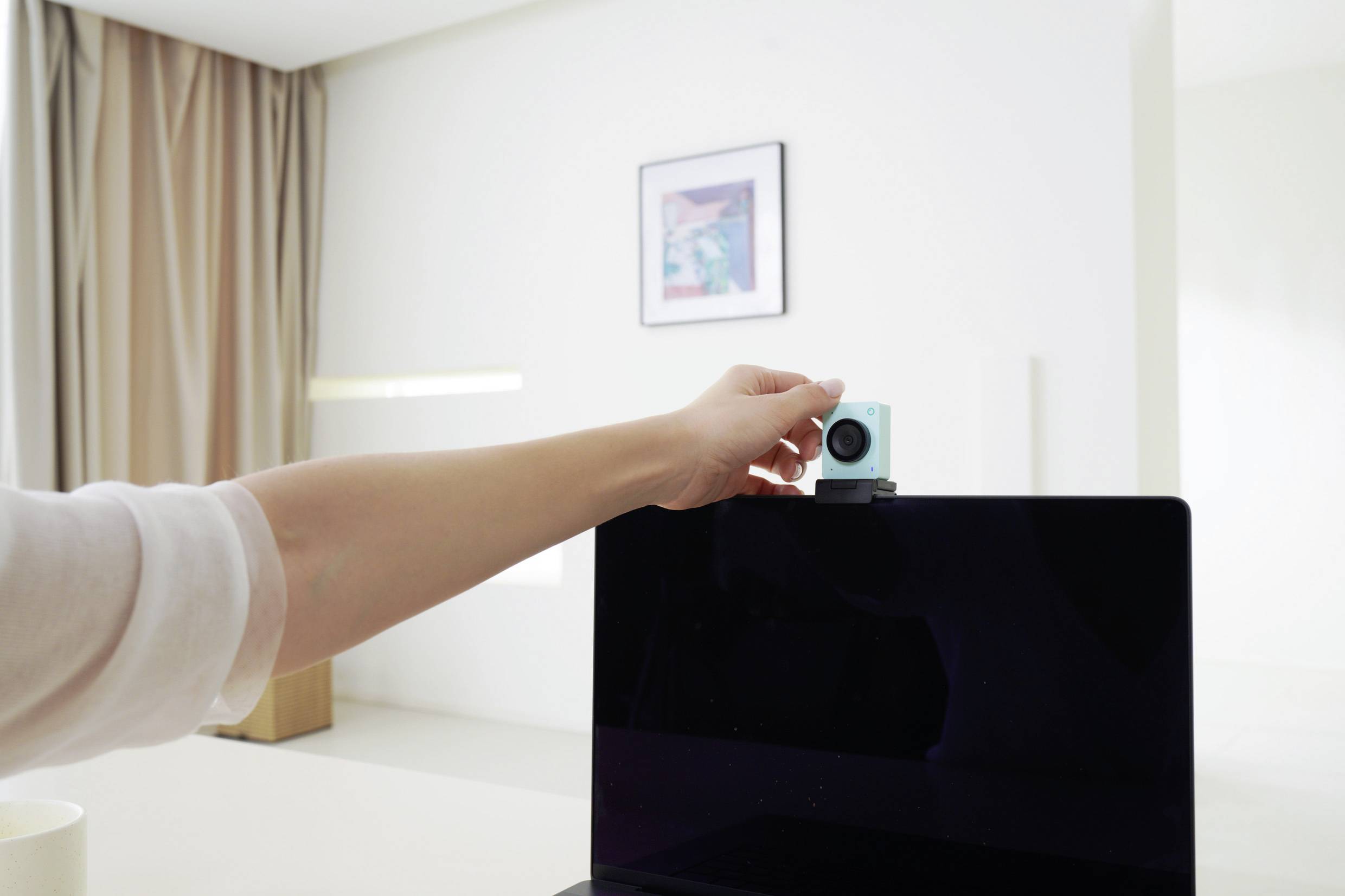 Une personne place une petite caméra sur l'écran d'un ordinateur portable dans une pièce lumineuse avec une image sur le mur en arrière-plan.