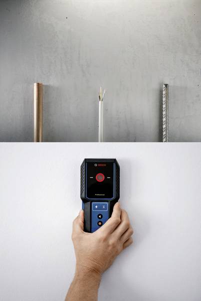Une main tient un appareil de mesure électronique contre un mur. Derrière, on aperçoit des tuyaux et des câbles qui traversent la paroi.