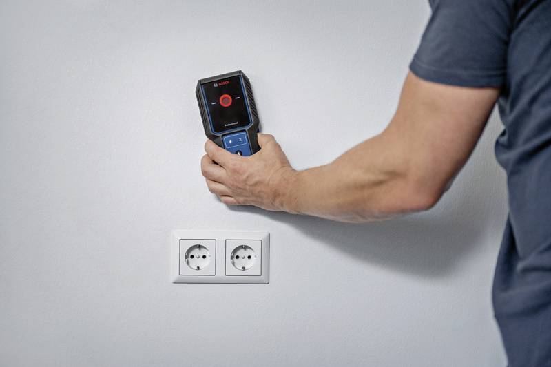 Une personne utilise un appareil électronique pour vérifier un mur via deux prises électriques sur un mur blanc.