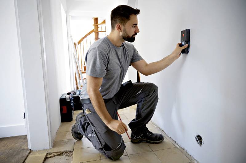 Un homme est à genoux dans un couloir et utilise un appareil numérique sur le mur pour localiser probablement des fils ou des tuyaux. Un escalier est visible en arrière-plan.