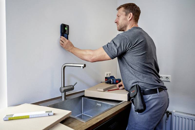 Un artisan utilise un appareil de mesure électronique sur un mur de cuisine. Des outils sont posés sur le plan de travail.