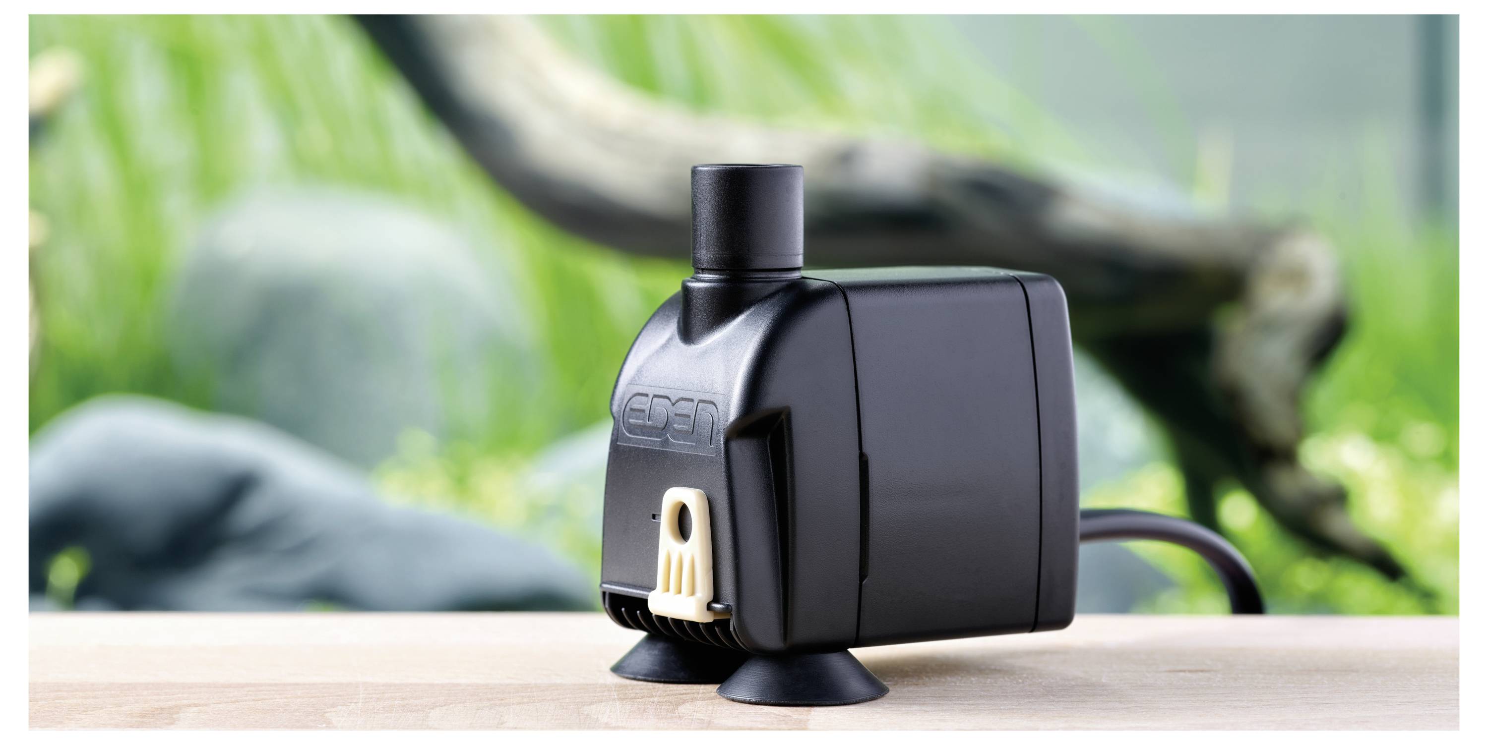 Pompe à eau noire d'aquarium sur une surface en bois, avec des plantes aquatiques vertes et du bois flotté flous en arrière-plan.