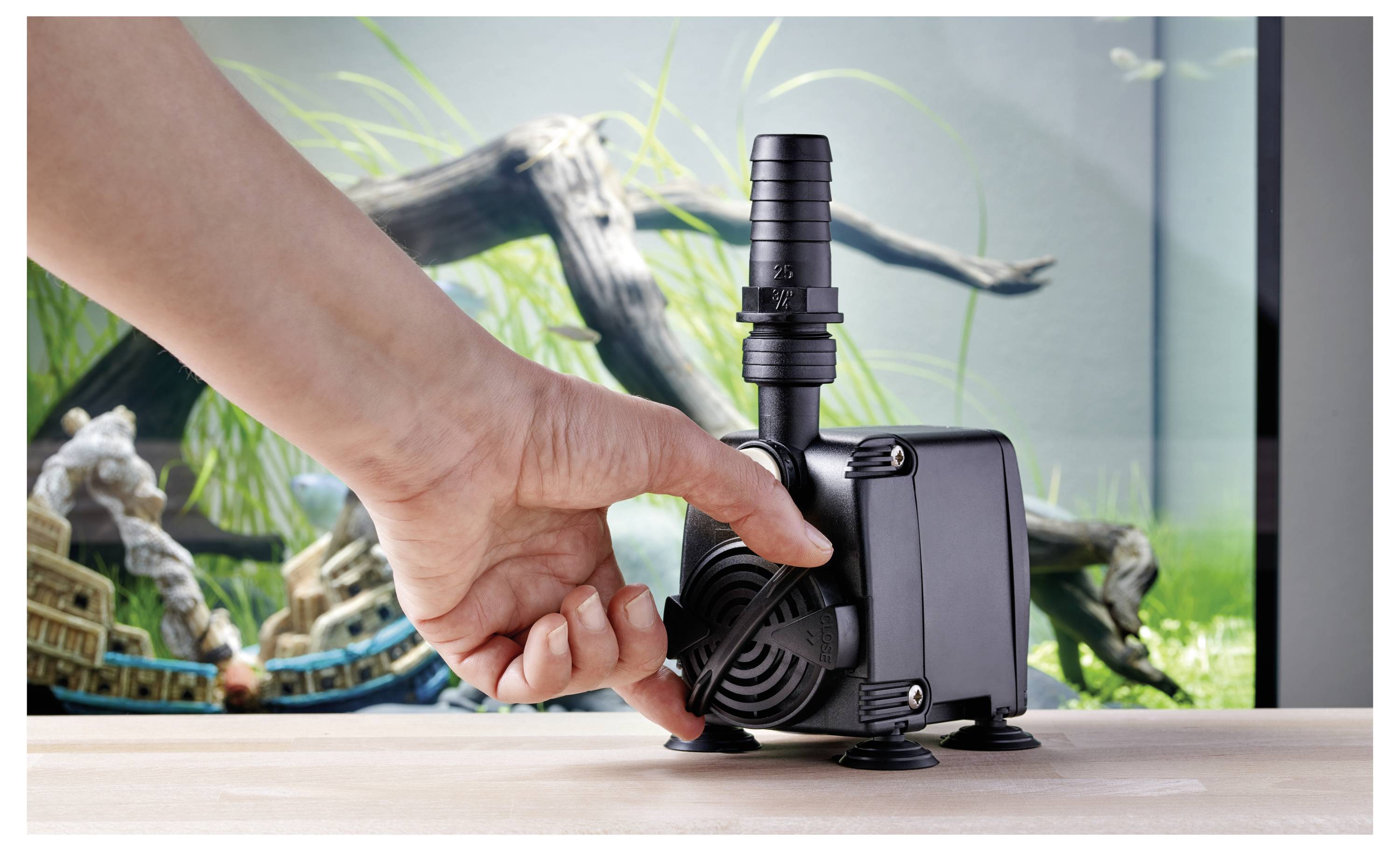 Une main ajuste une pompe de filtration noire pour aquarium avec des ventouses sur un plan de travail. Un aquarium avec de la verdure et des décorations est visible en arrière-plan.