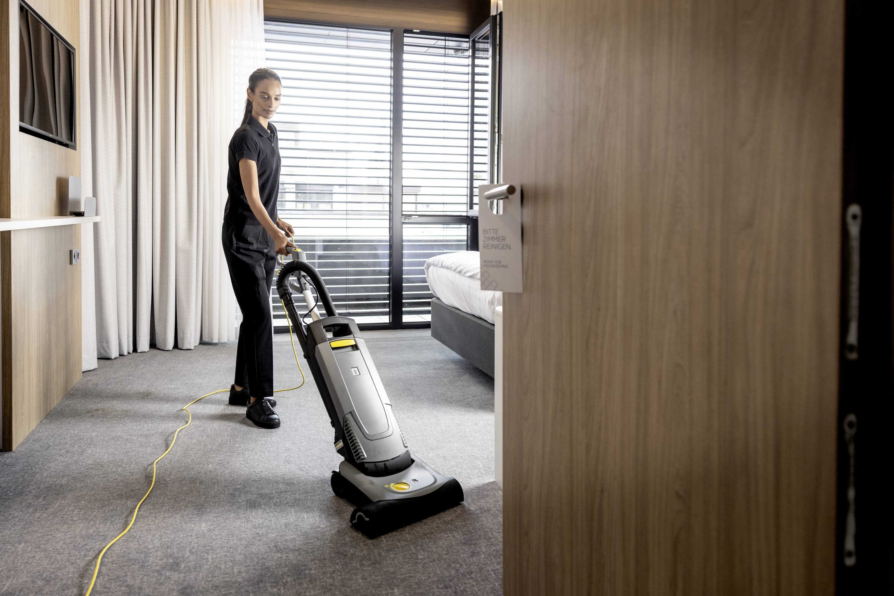 Une personne aspire le tapis avec un aspirateur dans une pièce moderne avec une grande fenêtre et des rideaux.