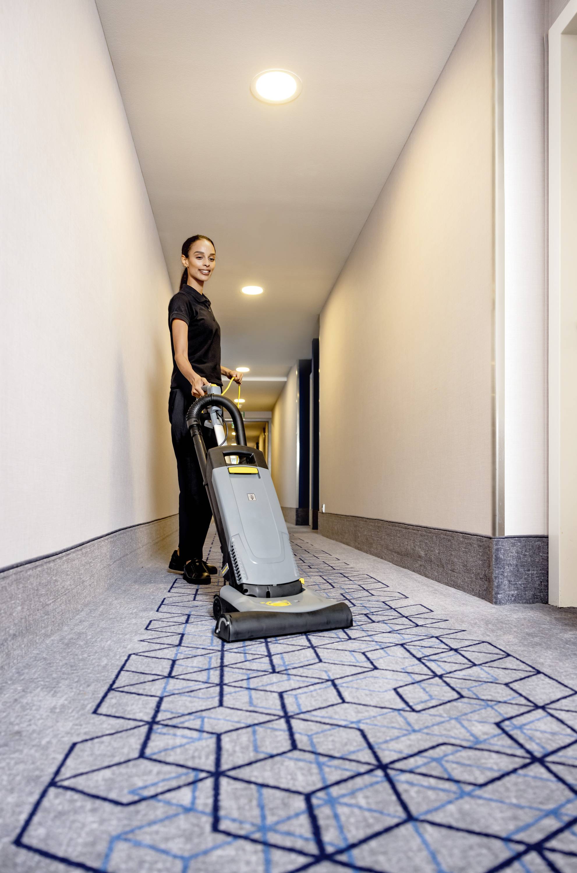 Une femme nettoie un couloir avec un aspirateur, dont le sol est recouvert d'un tapis au motif géométrique.