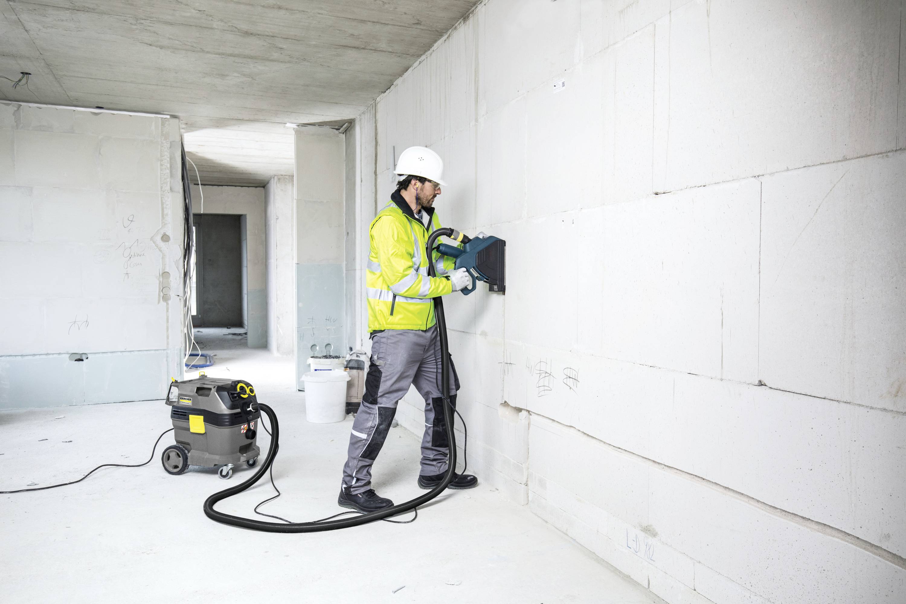 Un ouvrier portant des équipements de protection utilise un outil électrique dans une pièce en cours de rénovation. Un aspirateur est branché.