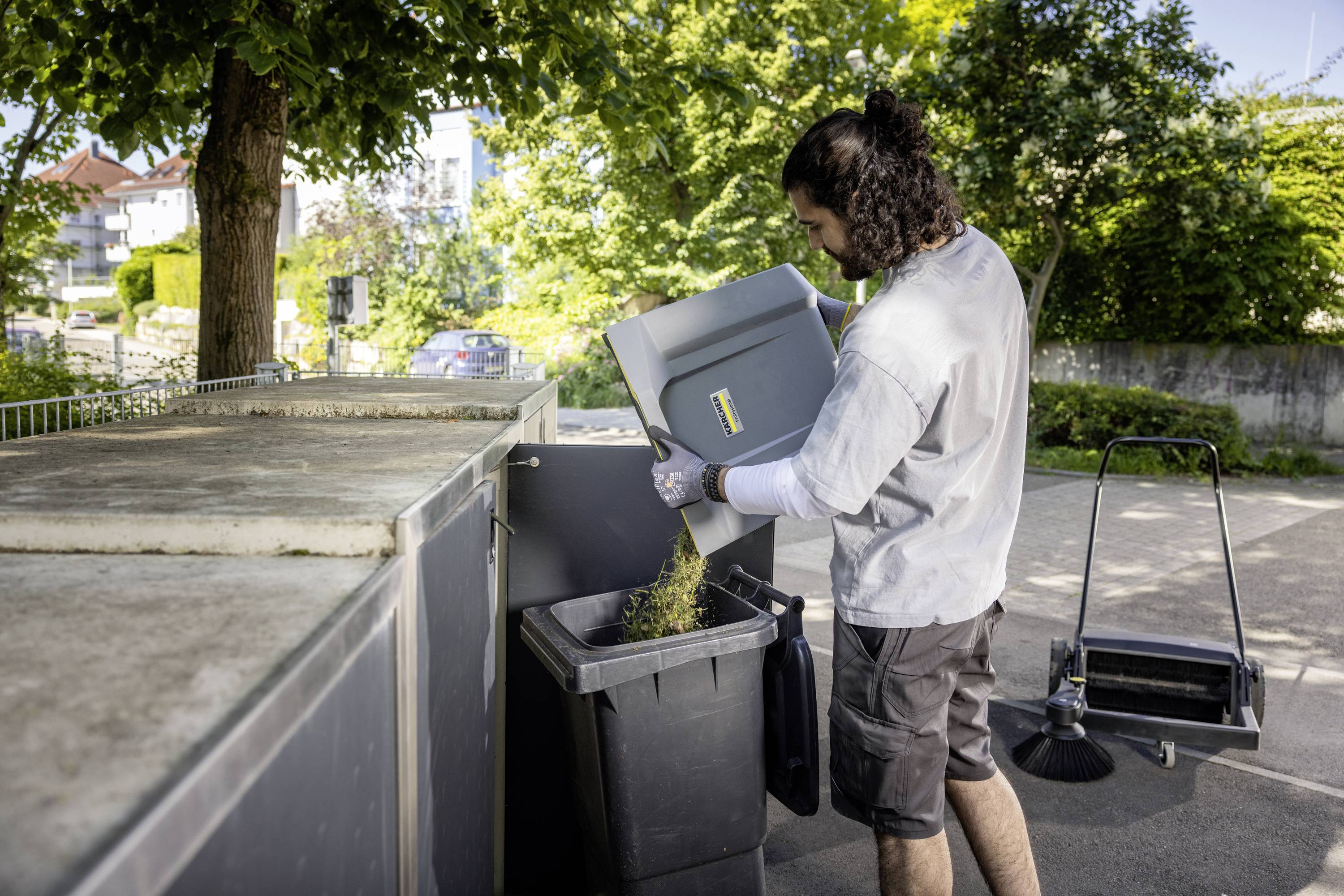 Une personne jette du carton dans un grand conteneur à déchets à l'extérieur. Des arbres et un trottoir sont visibles en arrière-plan.