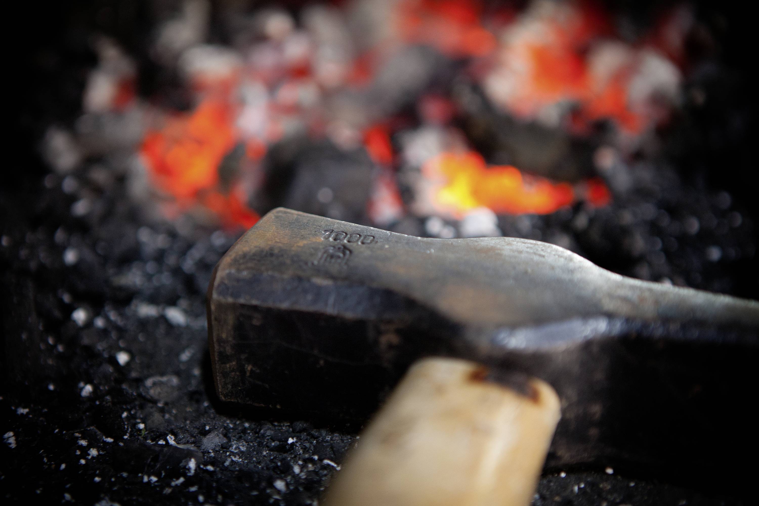 Un marteau est placé devant des charbons ardents. À l'arrière-plan, on aperçoit des flammes vives et des processus de fusion, ce qui suggère un processus de forge.