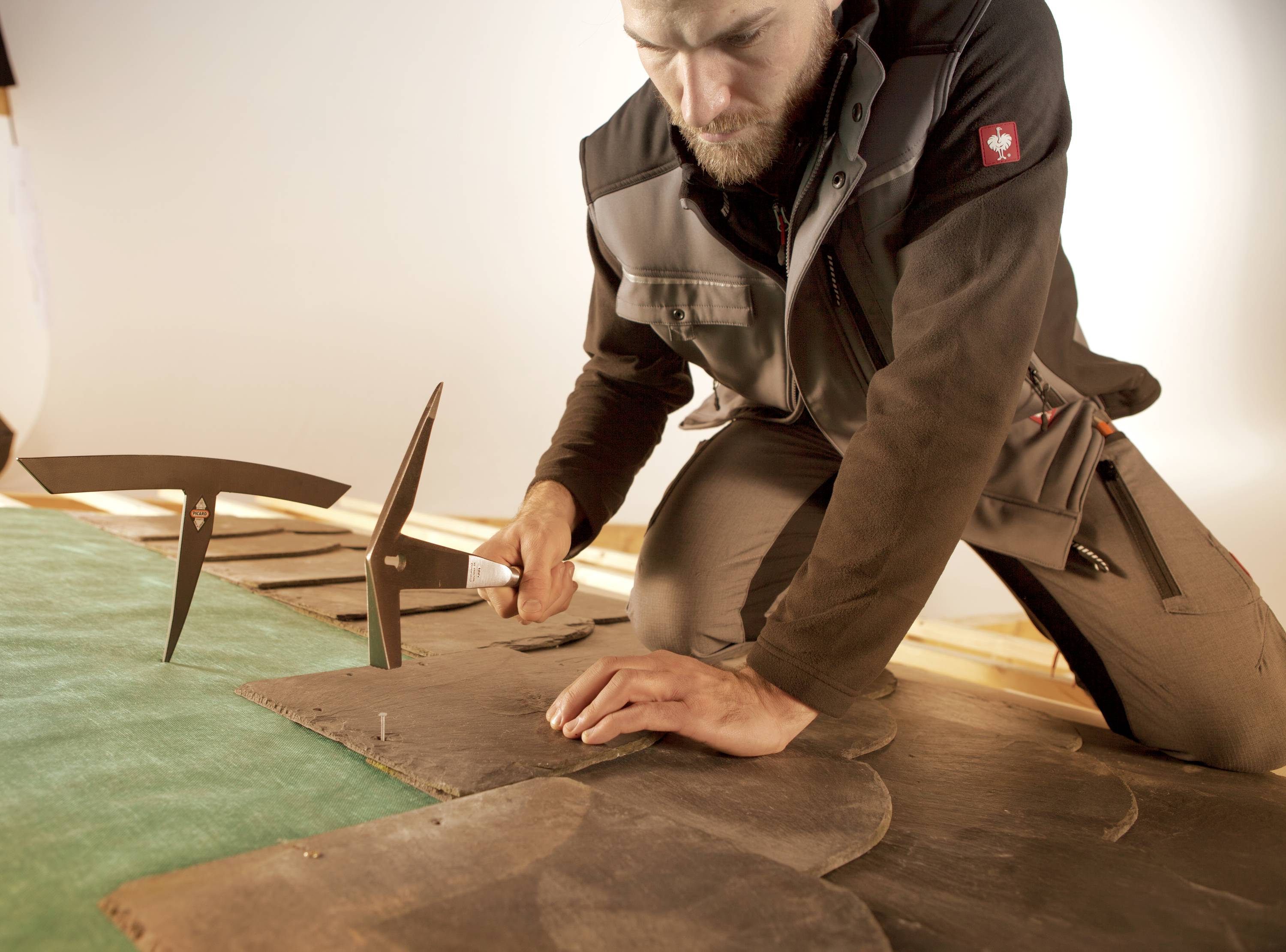 Un homme est agenouillé sur le sol et travaille avec des outils sur des bardeaux brun foncé. Il se concentre sur la précision du placement.