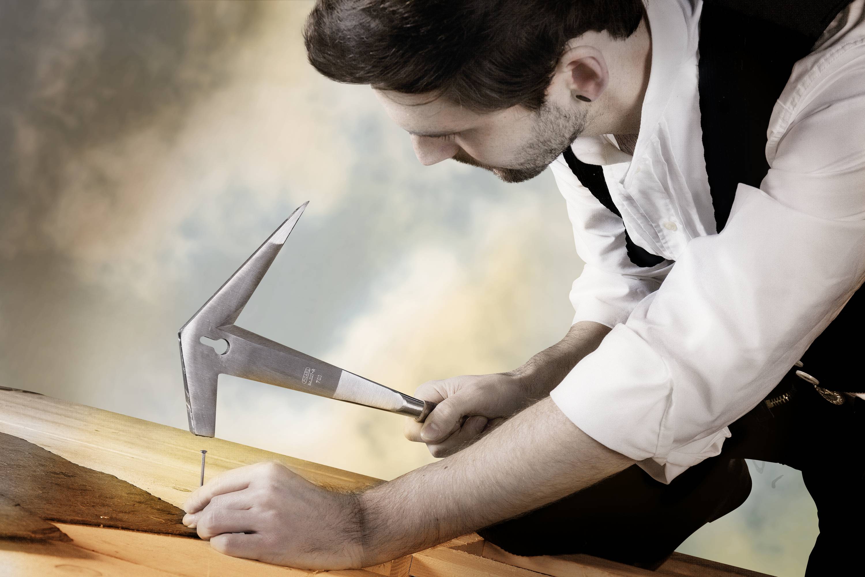 Un artisan vêtu de façon traditionnelle enfonce un clou dans une planche de bois en travaillant avec un marteau, le ciel étant visible en arrière-plan.
