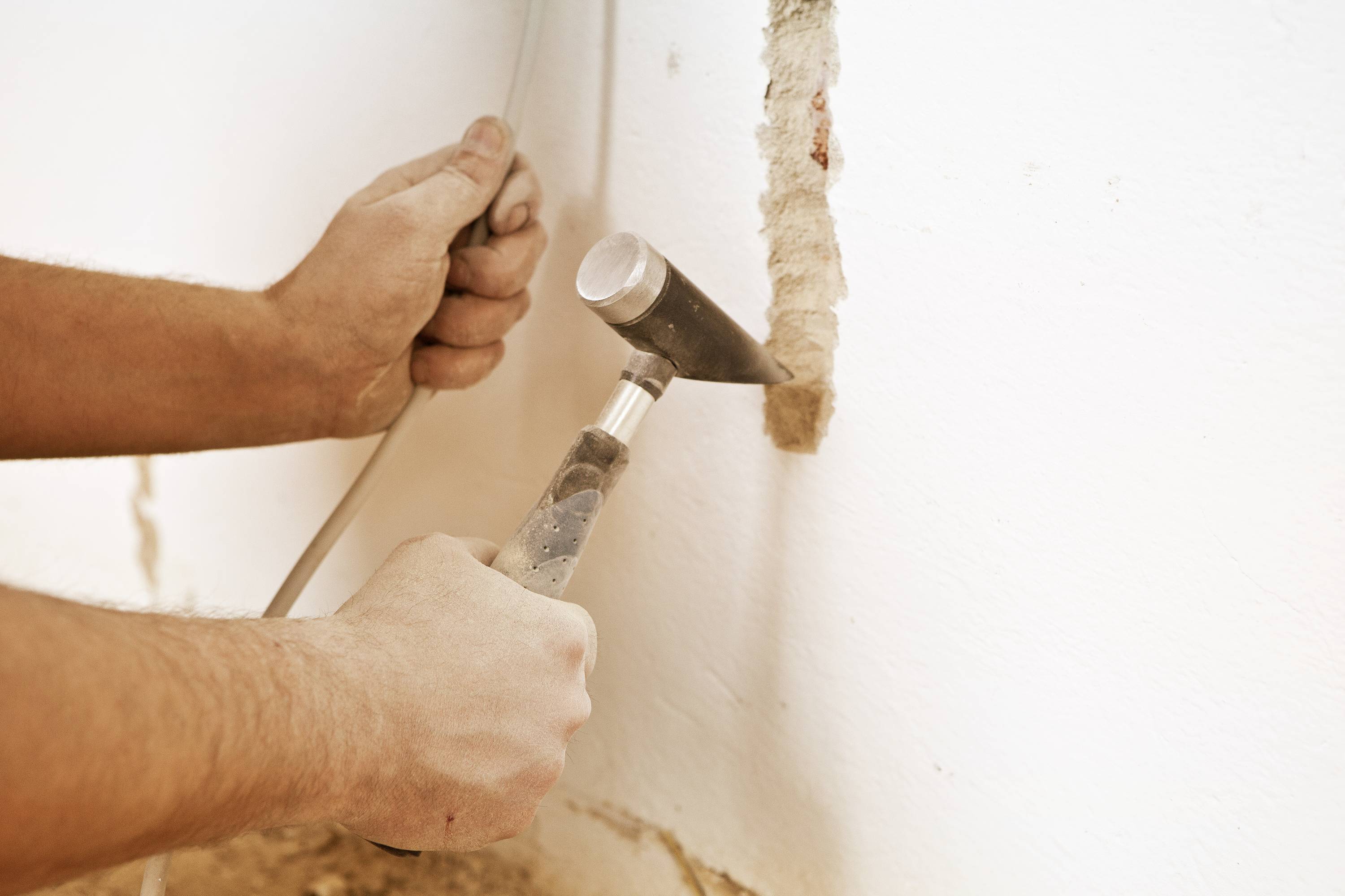 Une personne travaille avec un marteau et un burin sur un mur. Elle retire l'enduit pour dégager un câble situé en dessous.