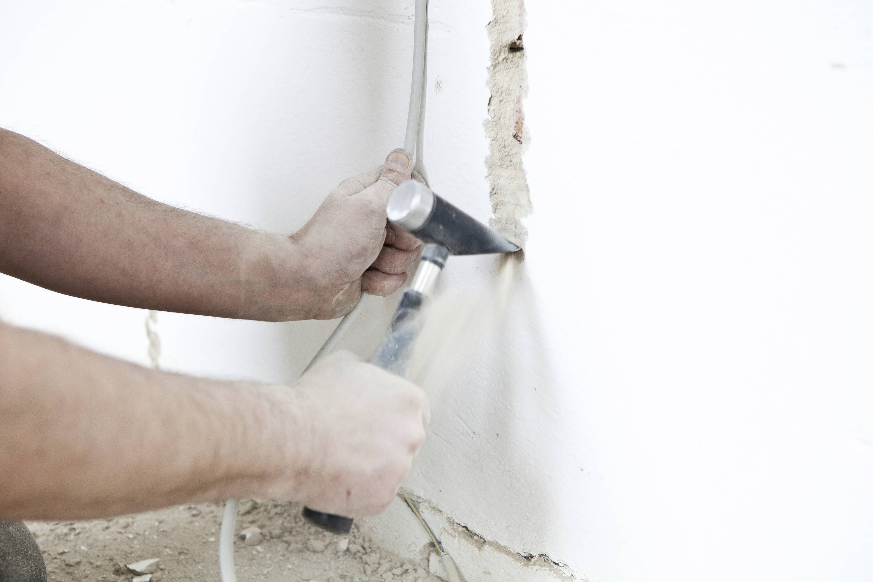 Une personne travaille avec un marteau et un burin pour encastrer un câble dans un mur. Le mur est blanc et légèrement endommagé.