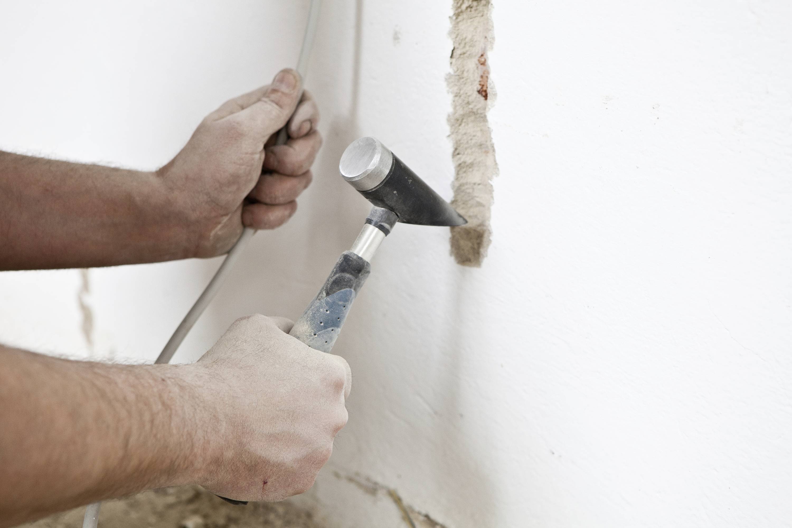 Une personne utilise un marteau et un burin pour percer un chemin de câbles dans un mur blanc. Les mains et le mur sont visibles.