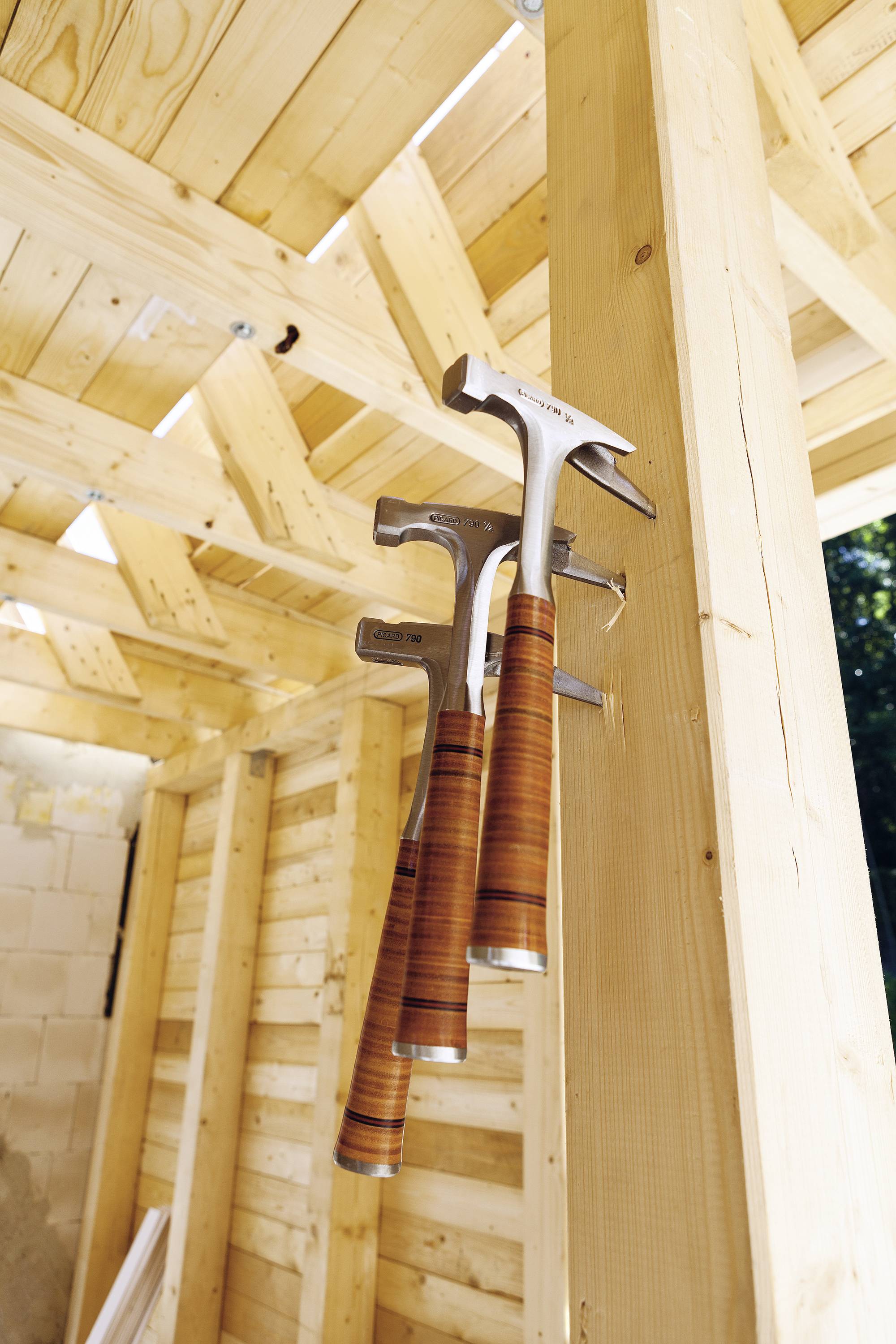 Trois marteaux sont suspendus au mur en bois d'une construction en bois semi-achevée. Le chantier est bien éclairé et montre des progrès dans la construction.