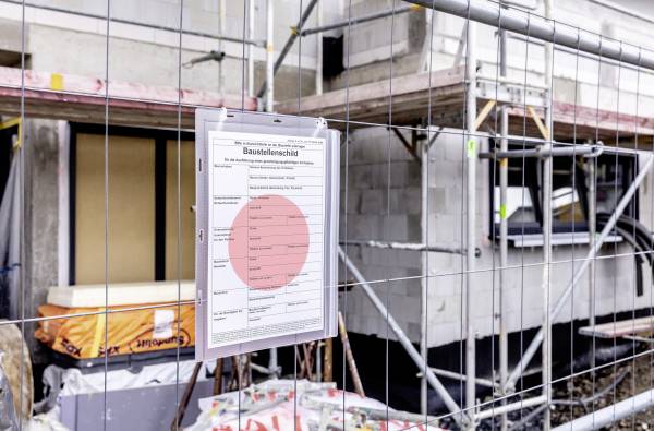Terrain de construction avec une clôture de sécurité et un panneau 'Chantier', affichant des informations de sécurité. Arrière-plan : structure en cours de construction d'un bâtiment.