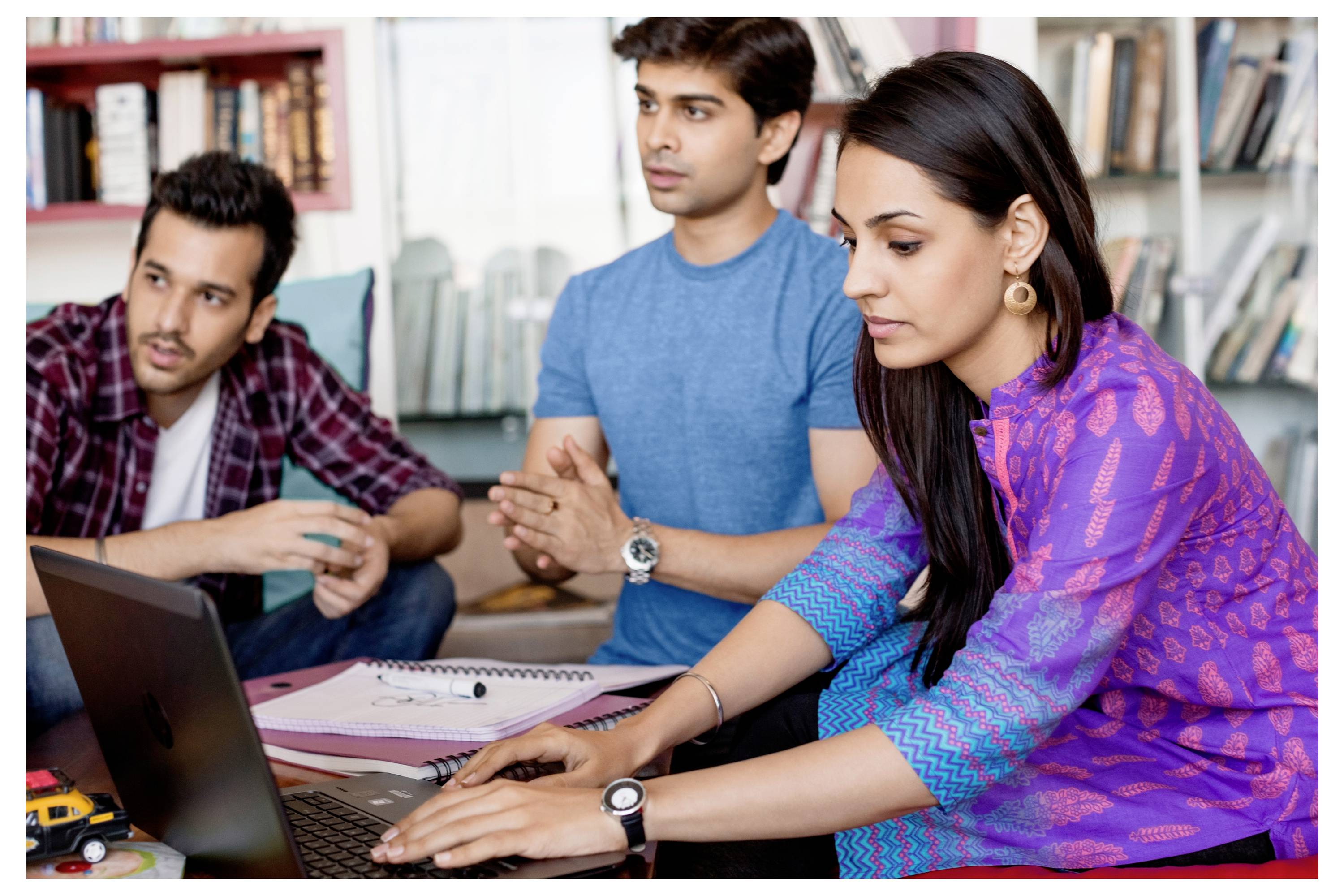 Trois personnes travaillent ensemble dans une pièce avec des livres en arrière-plan. Une personne tape sur un ordinateur portable pendant que les autres regardent.