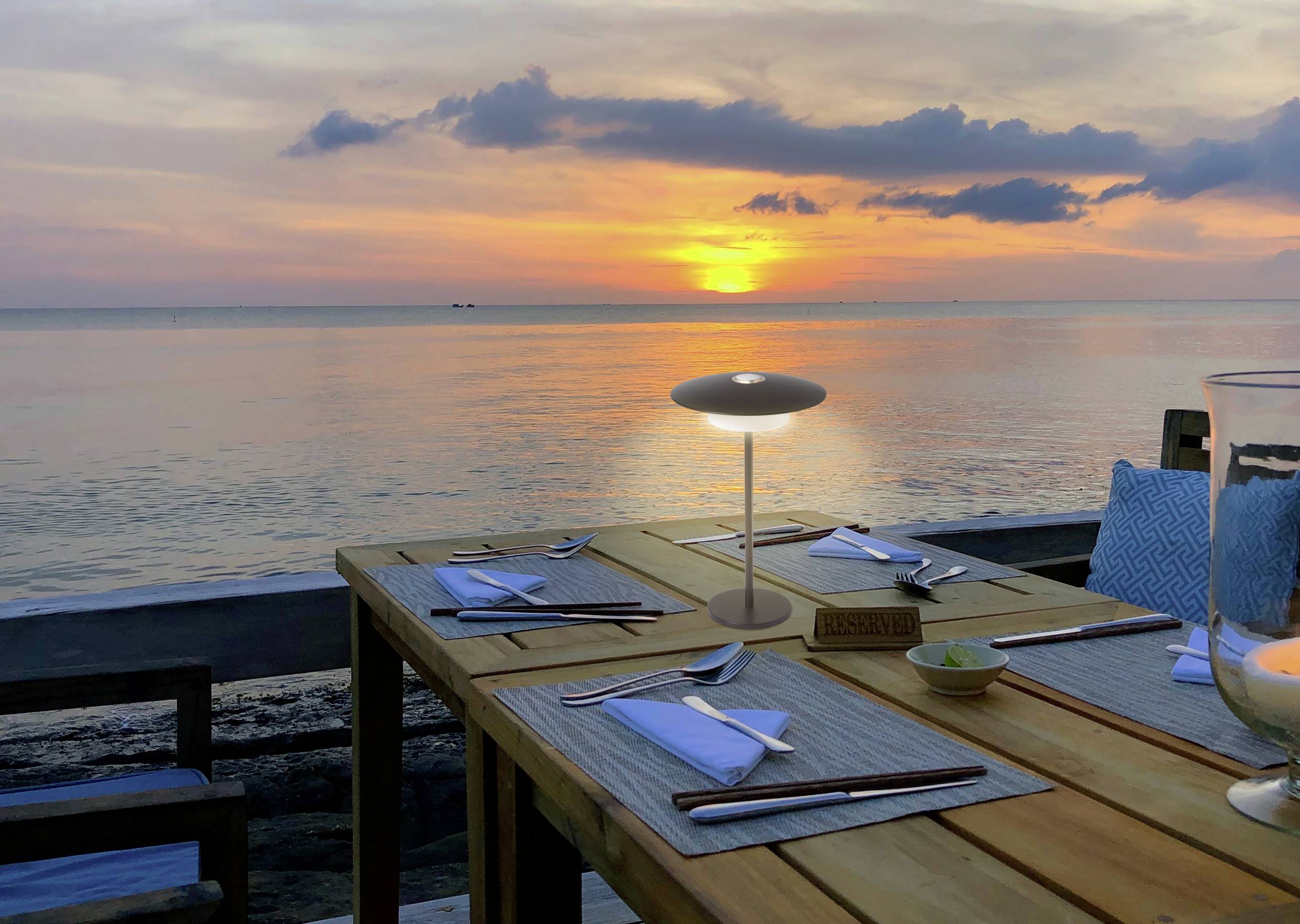 Table à manger dressée pour deux personnes avec des couverts et des serviettes, donnant sur un coucher de soleil océanique serein avec une lueur orange se reflétant sur l'eau.