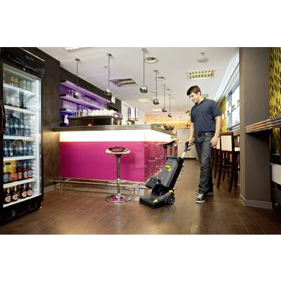 Un homme nettoie le sol d'un bar moderne à l'aide d'un appareil de nettoyage. À l'arrière-plan, on peut voir des sièges colorés et un comptoir éclairé.