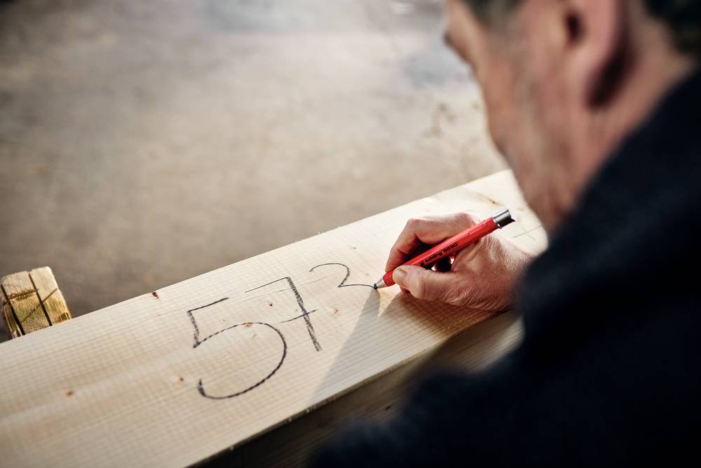 Une personne écrit le chiffre '57' avec un stylo rouge sur un morceau de bois. La scène suggère une activité artisanale.