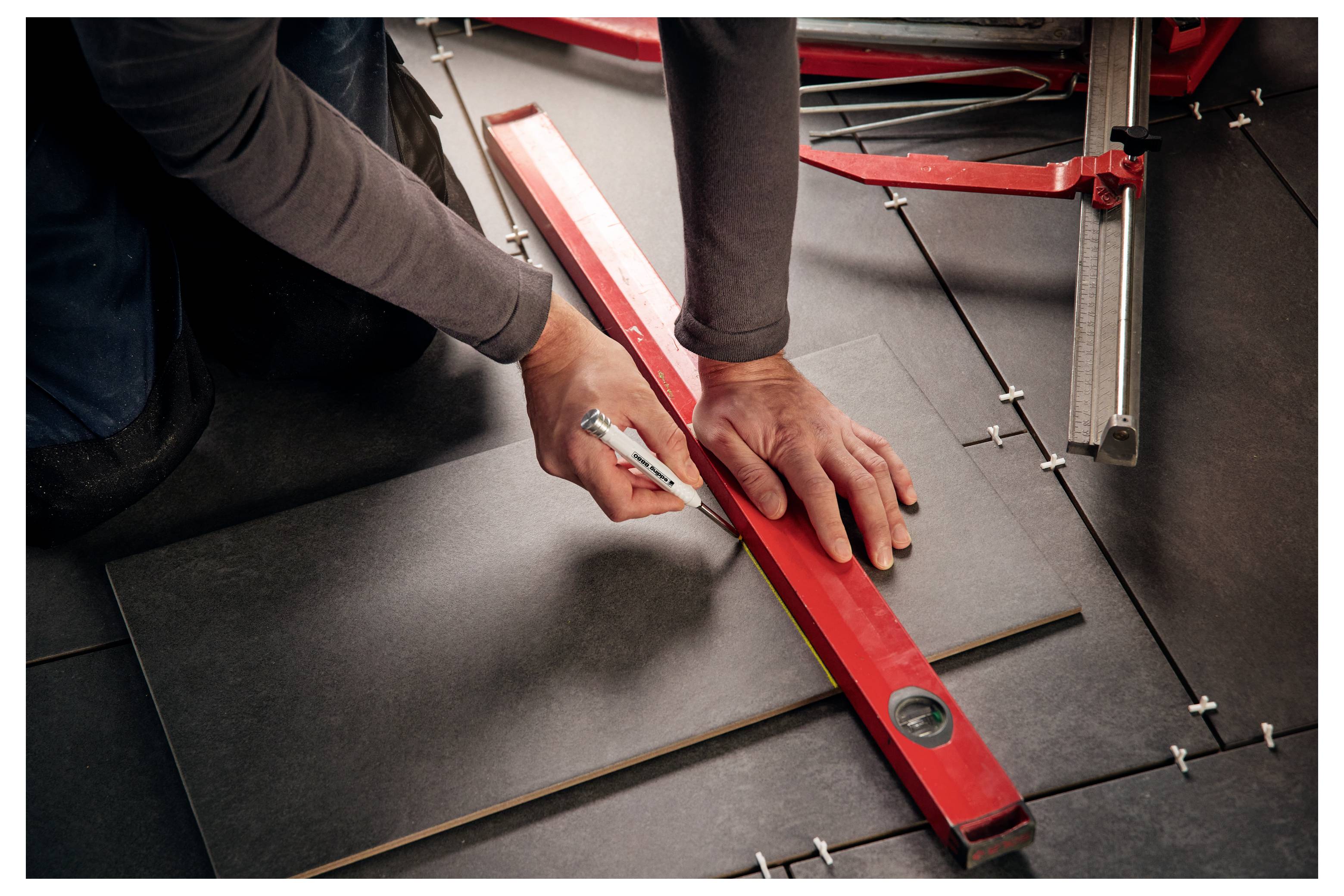 Une personne utilisant un couteau utilitaire pour couper une dalle de sol noire le long d'une règle droite rouge lors de l'installation.