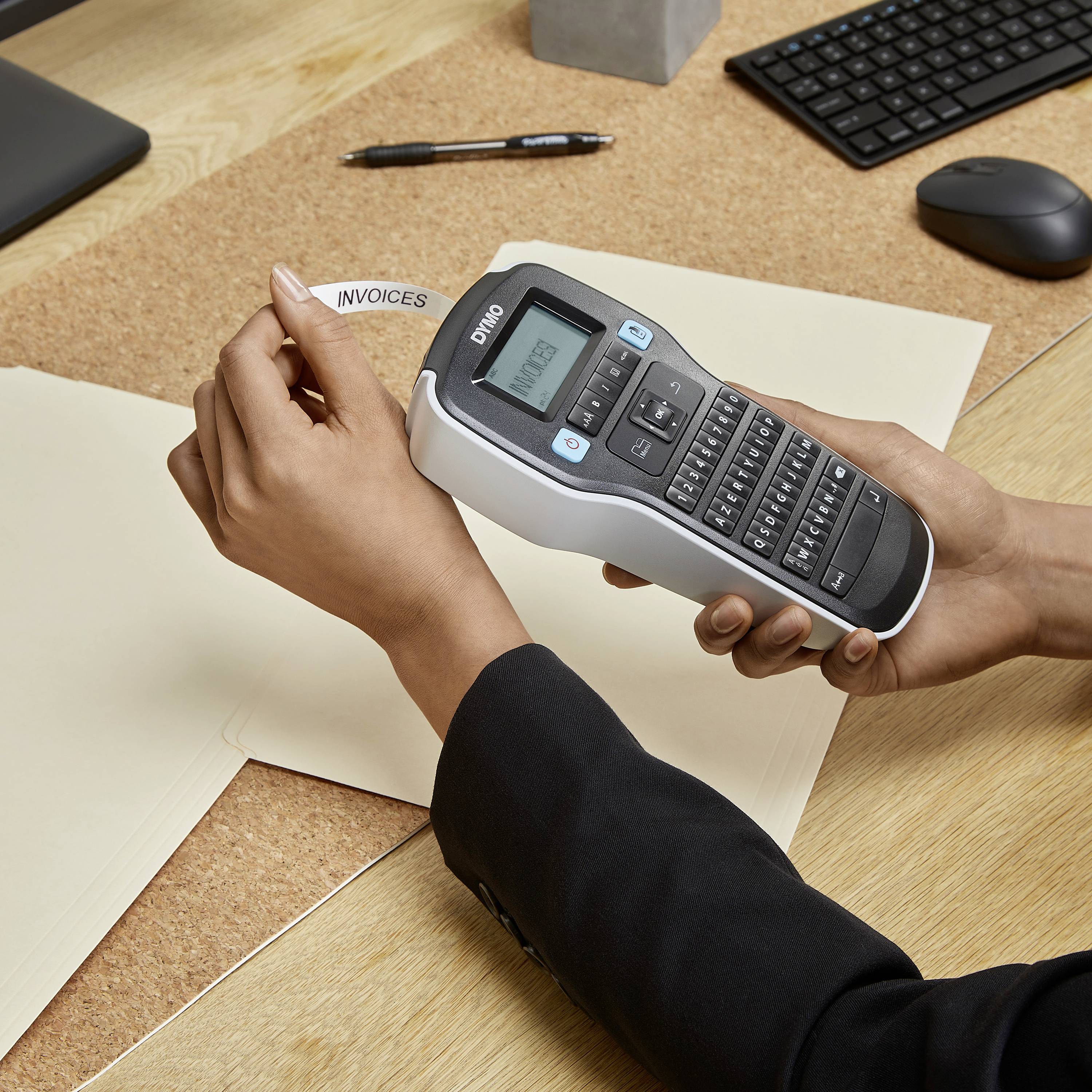 Une main tient un étiqueteuse qui imprime une étiquette portant l'inscription 'INVOICES'. Sur la table sont posés des enveloppes, un ordinateur portable et une souris.