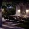 Une terrasse accueillante de nuit, éclairée par des appliques murales. Une table à manger avec de la vaisselle et quatre chaises occupe le centre, entourée de plantes et d'un canapé.