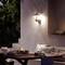 Un salon extérieur avec une table dressée pour un dîner. Une lampe murale diffuse une lumière chaleureuse, entourée d'arbres.