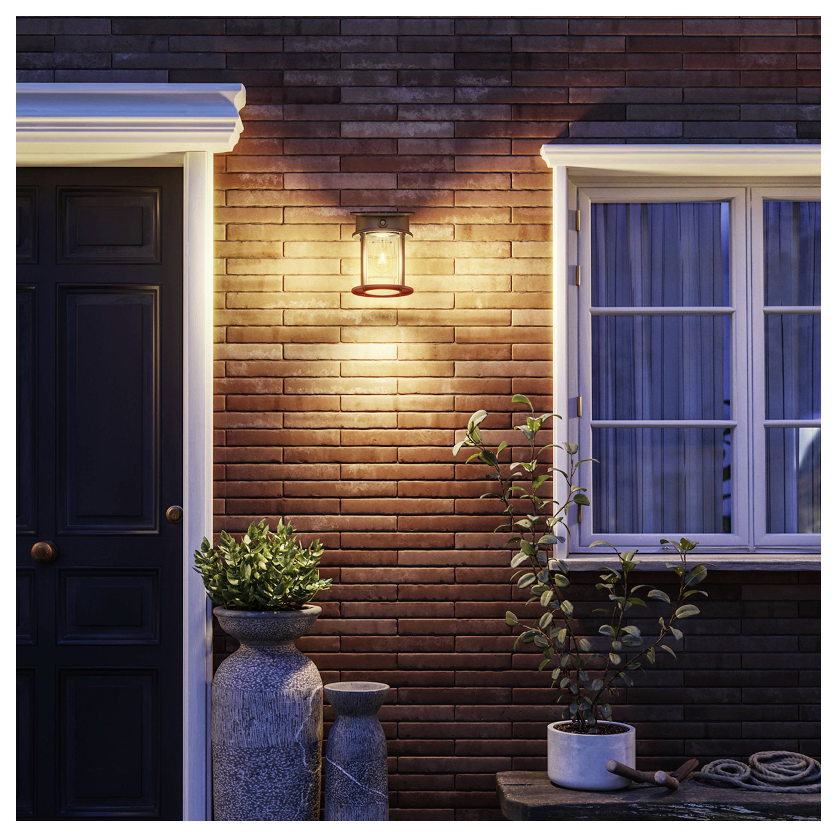 Mur extérieur d'une maison en brique au crépuscule avec une lanterne illuminée, une porte d'entrée sombre, une fenêtre et des plantes en pot près de l'entrée.