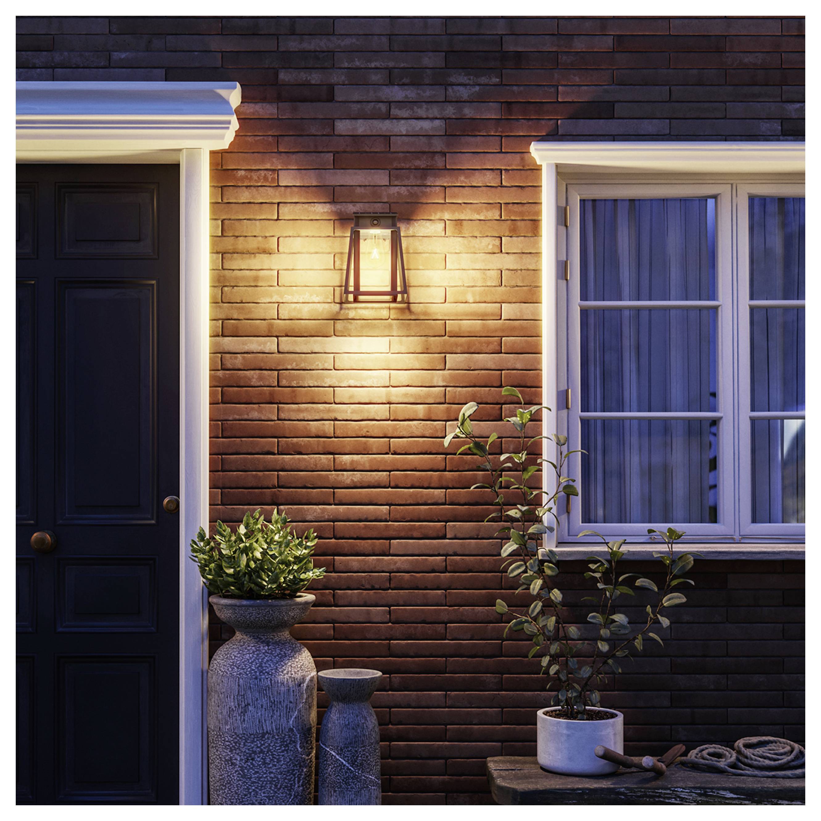 Extérieur d'une maison en brique au crépuscule avec un applique murale éclairée, une porte d'entrée sombre, une fenêtre et des plantes en pot sur un porche.