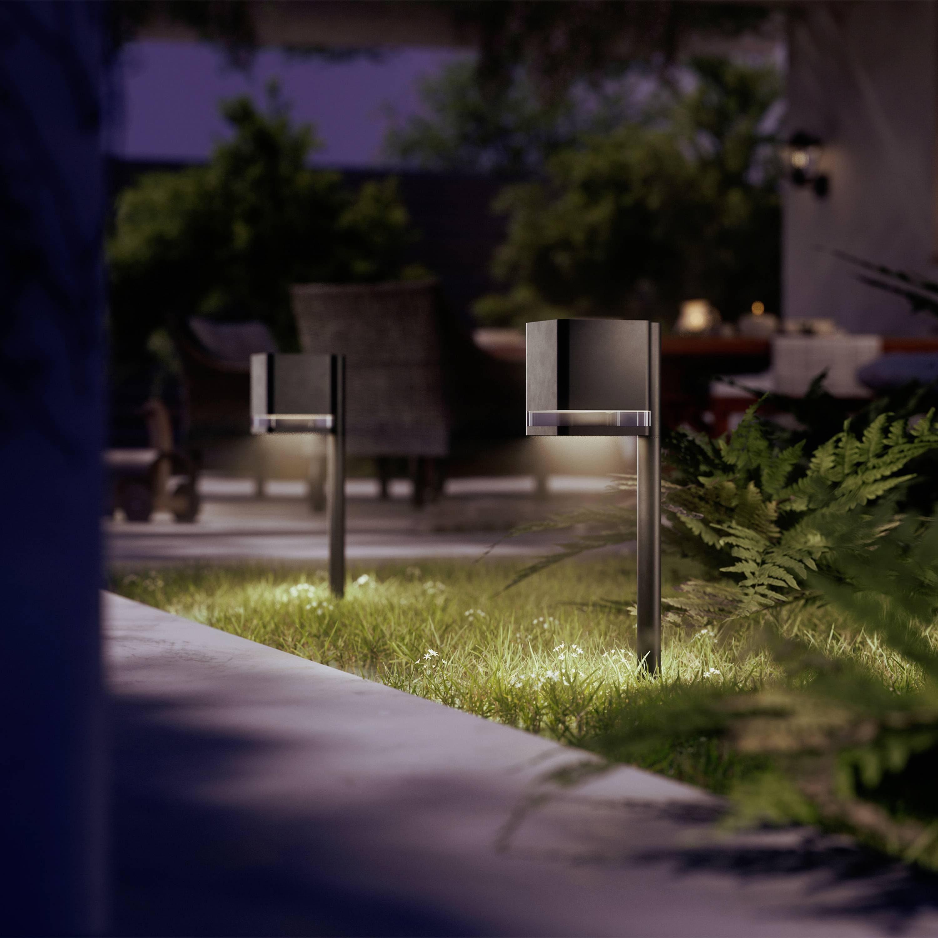 Deux luminaires modernes éclairent un espace de jardin soigné au crépuscule, entourés de plantes avec un arrière-plan flou.