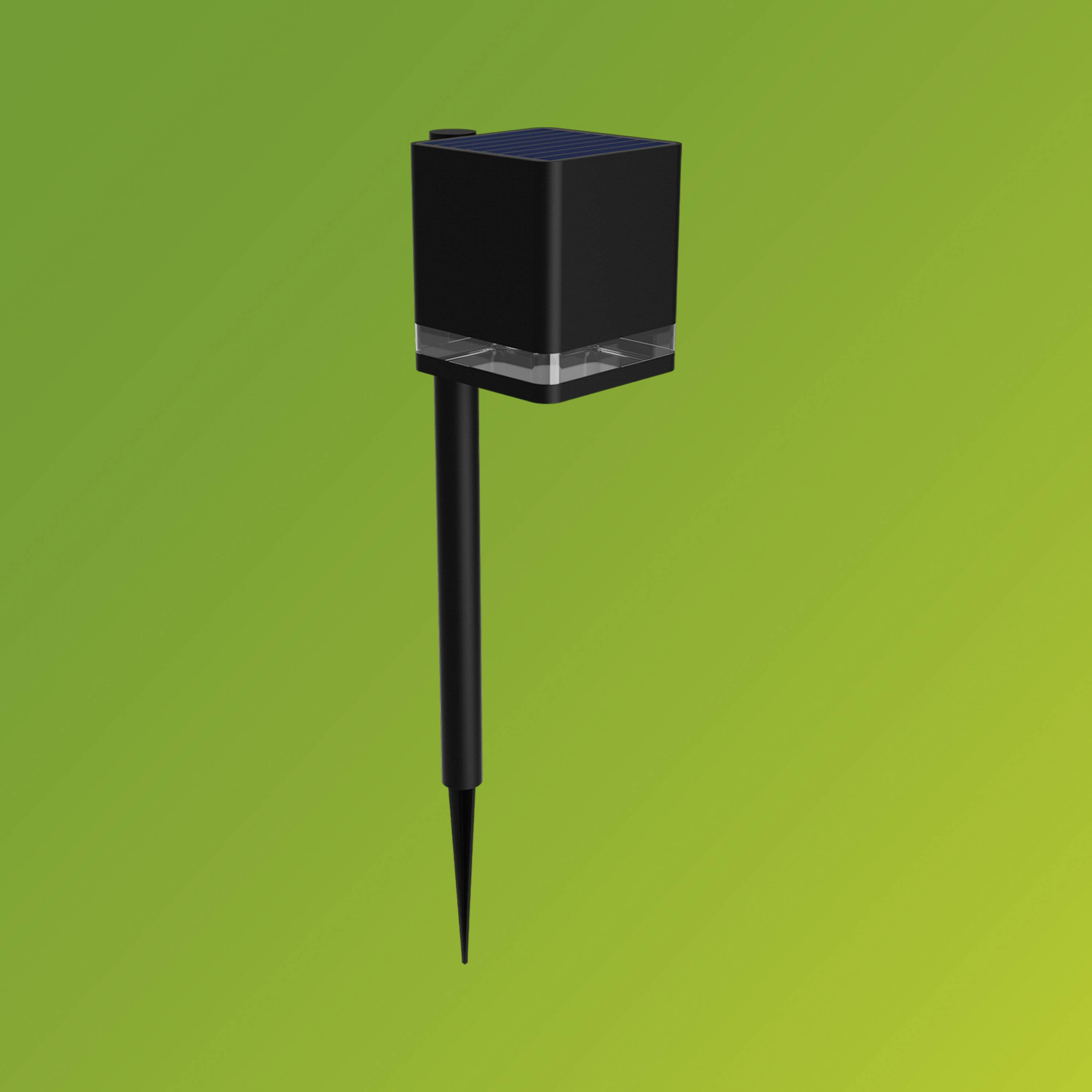 Un appareil noir, cubique, avec un piquet de terre sur un fond vert, probablement un éclairage de jardin solaire.