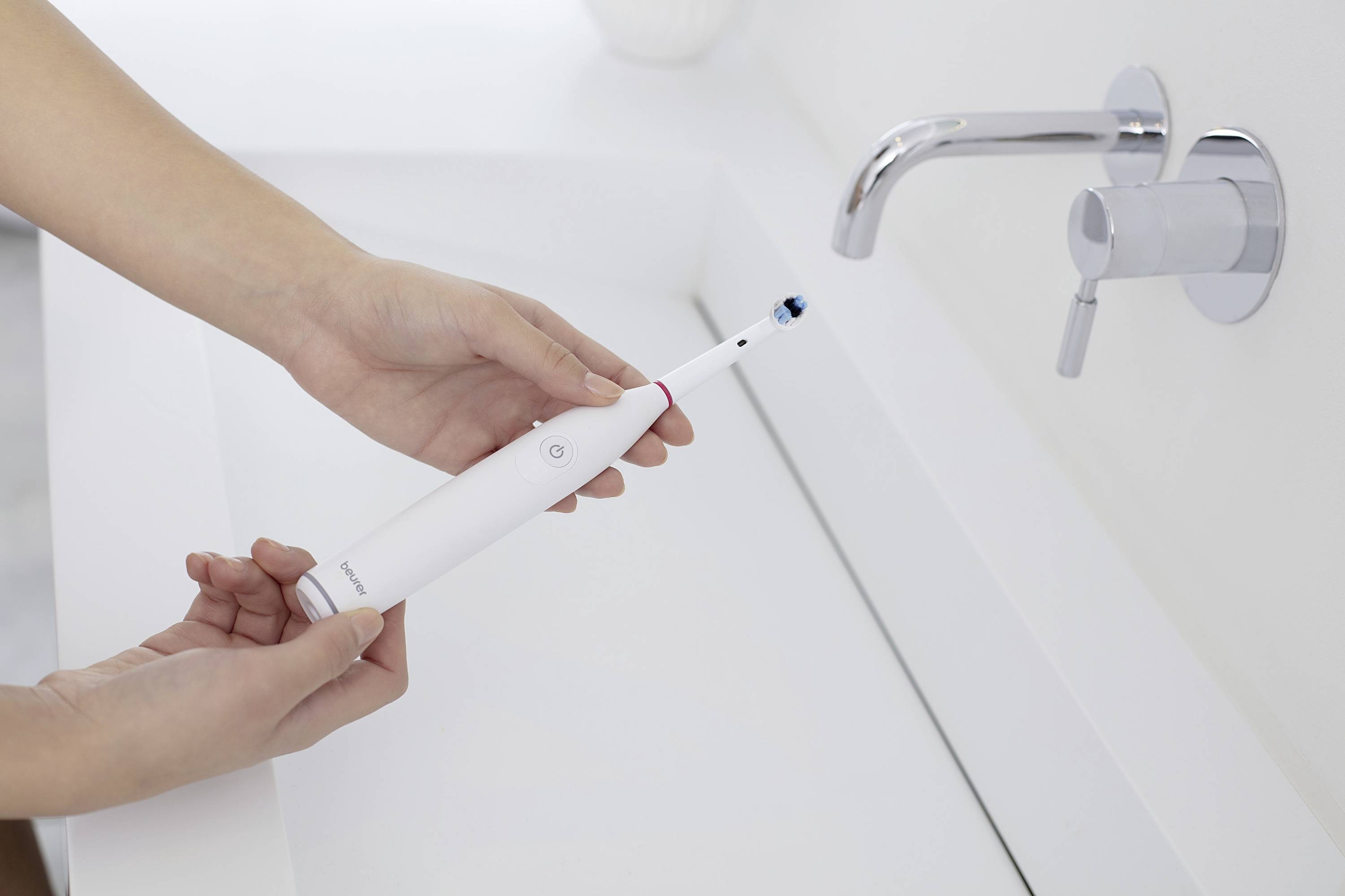 Une personne tient une brosse à dents électrique au-dessus d'un lavabo, prête à être utilisée. Un robinet est à proximité.