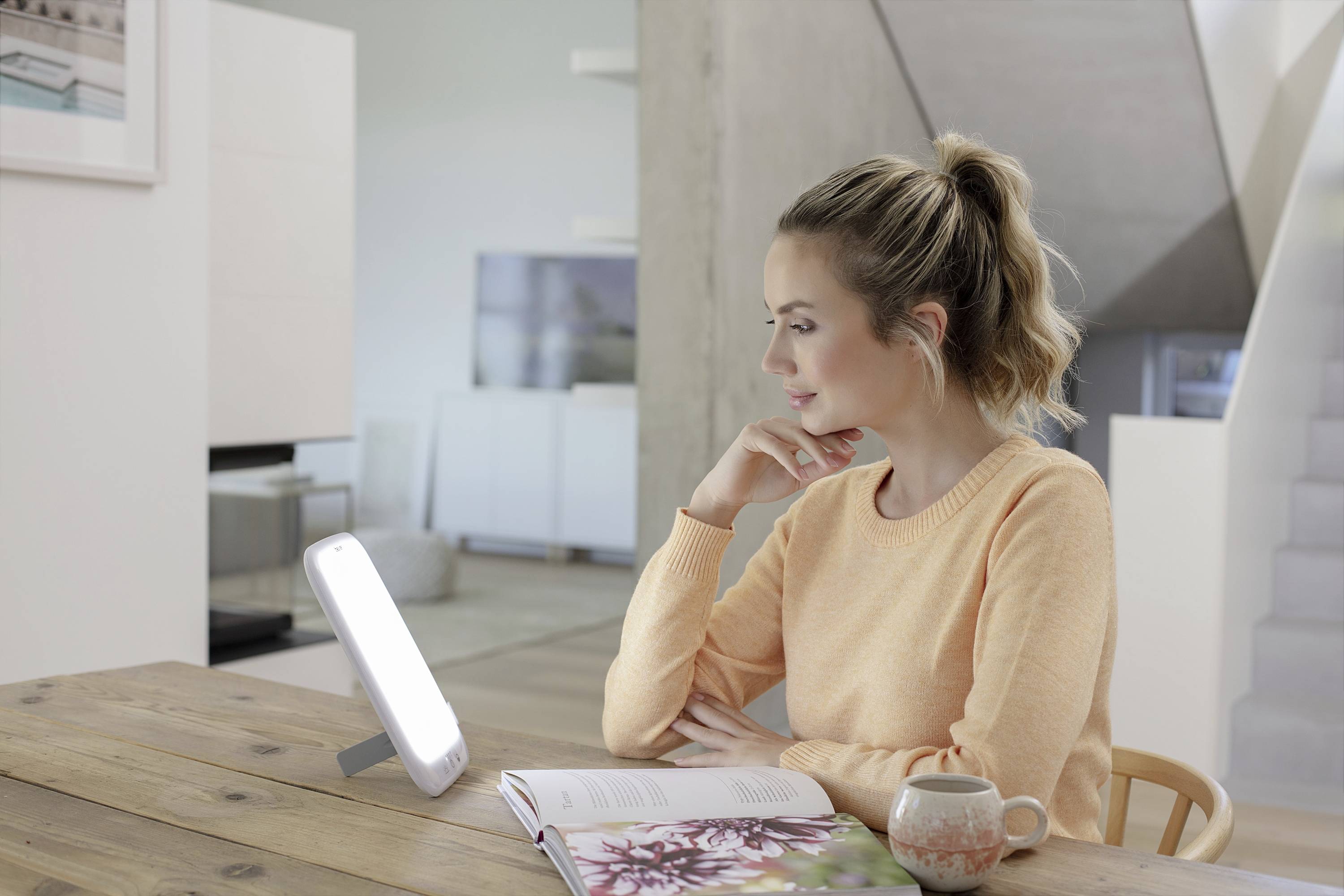 Une femme est assise à une table en bois, regardant pensivement une lampe de luminothérapie et tenant une tasse près d'un livre ouvert.