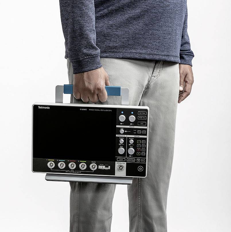 Une personne tient un oscilloscope portable avec une poignée à la main, prête à être utilisée. L'appareil dispose de divers boutons et affichages.