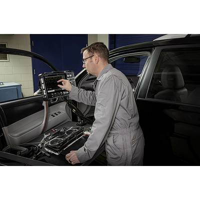 Un mécanicien en combinaison grise vérifie l'électronique d'une voiture à l'aide d'un outil de diagnostic à l'intérieur d'un atelier.