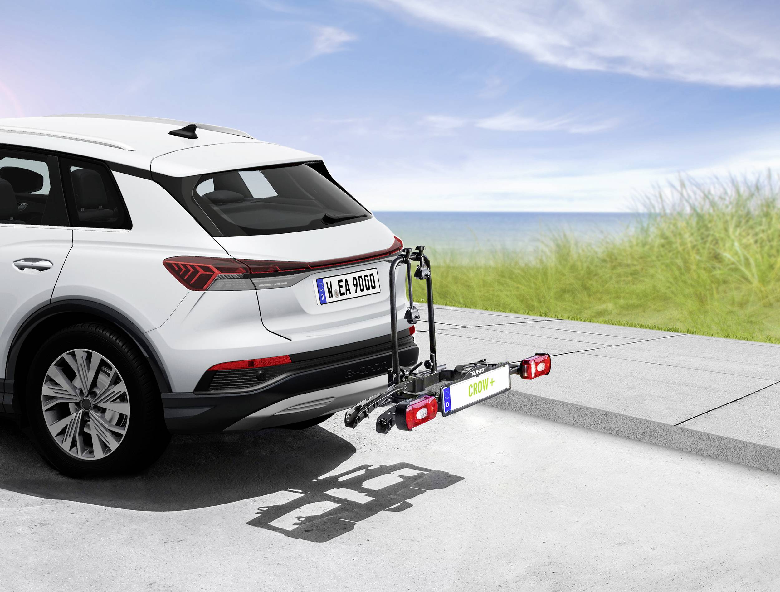 Un SUV blanc avec un porte-vélos à l'arrière, garé sur un chemin avec vue sur mer et une rive herbeuse en arrière-plan.