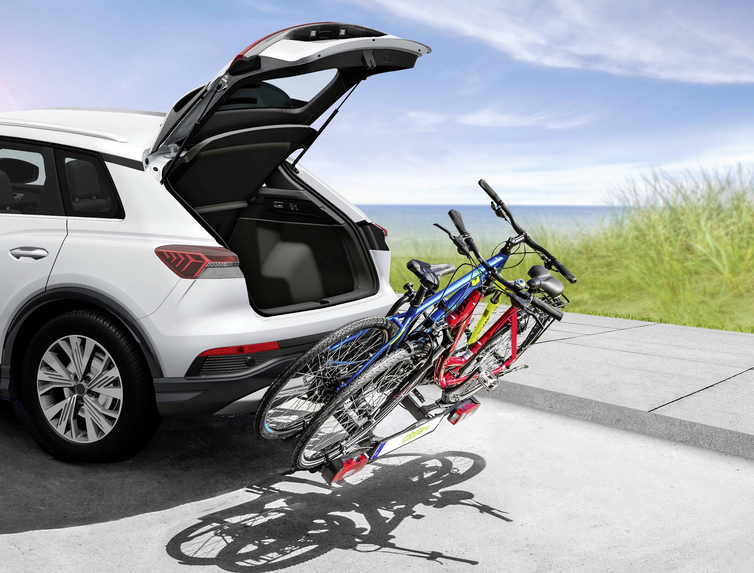 Une voiture blanche avec le coffre ouvert est garée sur la plage. Deux vélos sont montés sur le porte-vélos.