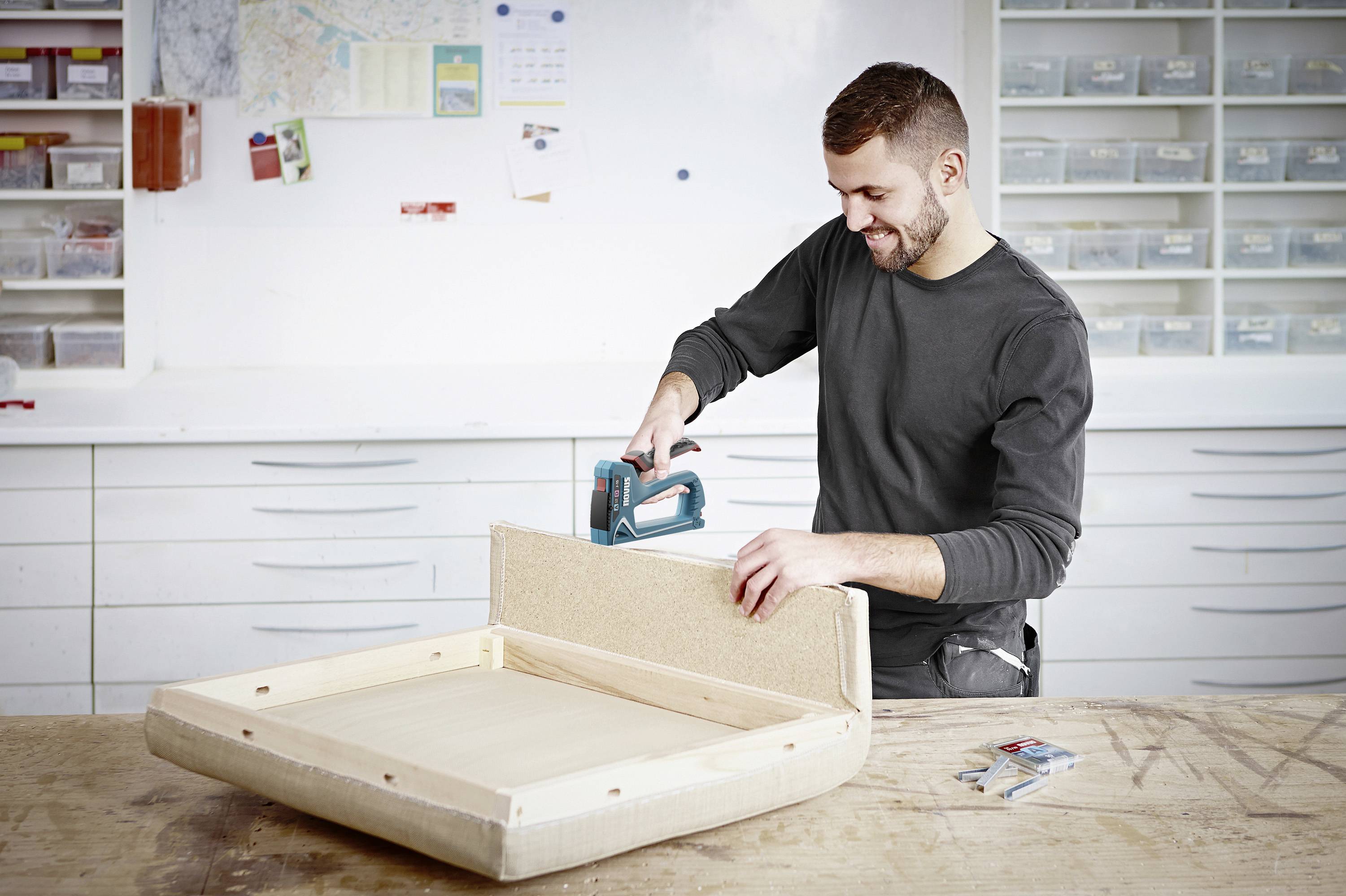 Un homme utilise un agrafeur électrique pour fixer un coussin sur un cadre en bois dans un atelier. À l'arrière-plan, on aperçoit des tiroirs d'atelier et des outils.