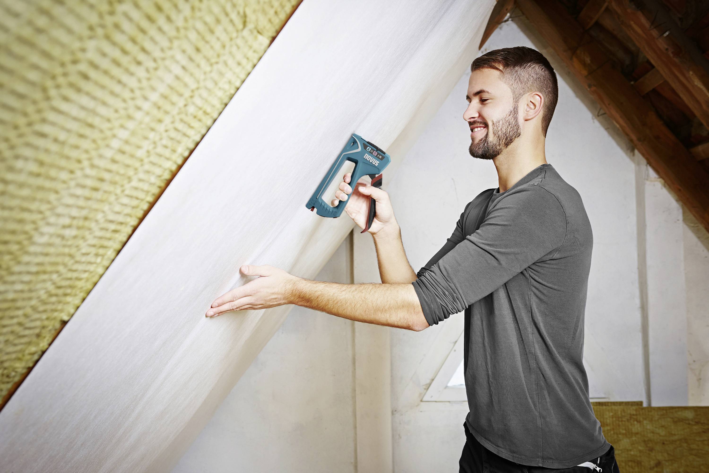 Un homme fixe un papier peint blanc sur un mur de toit incliné à l'aide d'un agrafeur électrique.
