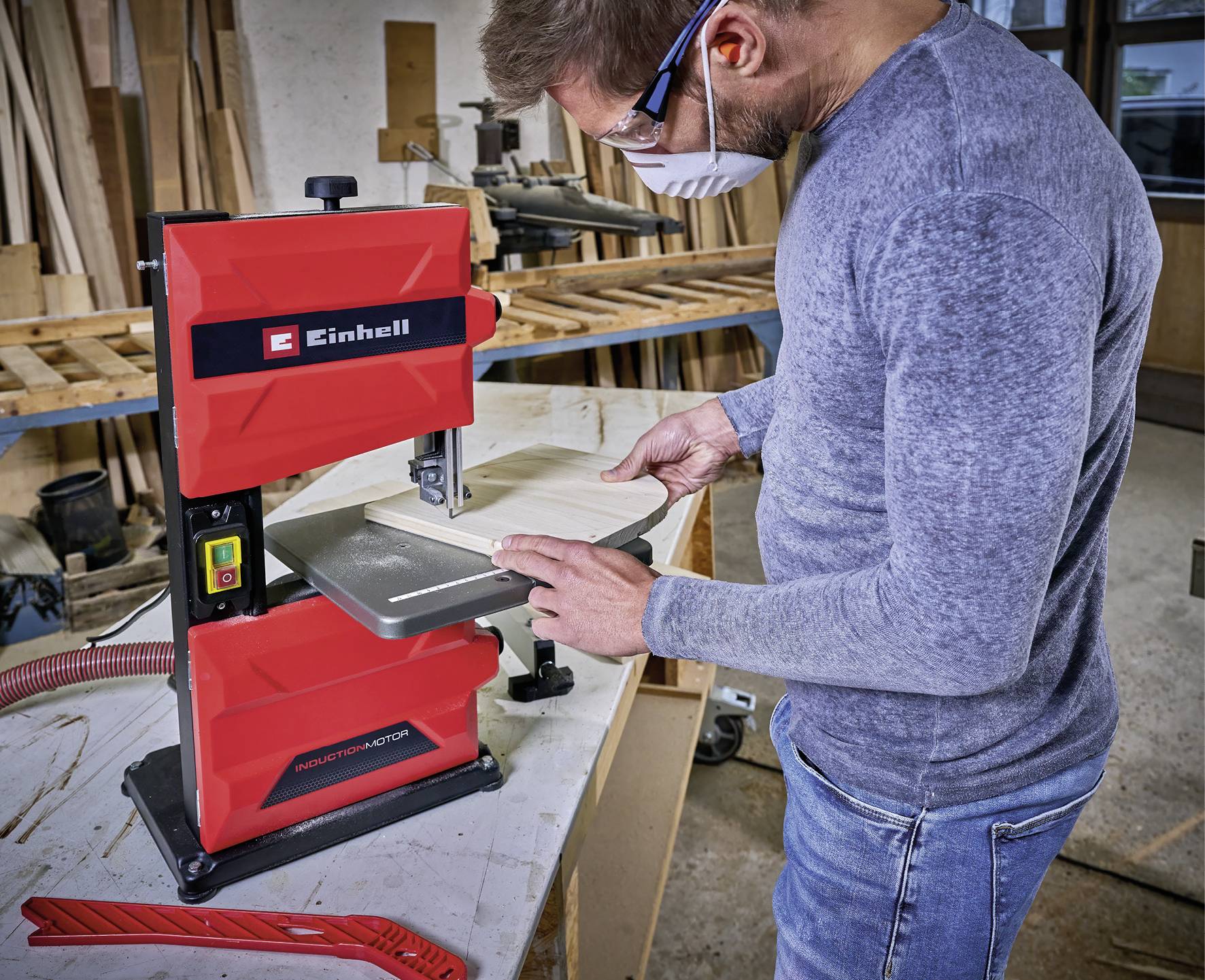 Un homme travaille avec une scie à ruban rouge dans un atelier. Il porte des lunettes de protection et un masque, et coupe un morceau de bois.